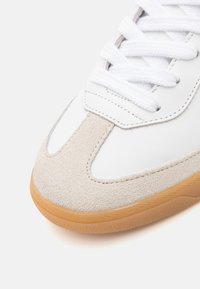 Gros plan sur la pointe d'une basket blanche avec un empiècement en daim beige, des lacets blancs et une semelle en caoutchouc naturel sur un fond clair.