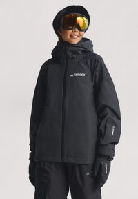 Person klädd i svart vinterutrustning för skidåkning med handskar, pälsmössa och reflekterande skidglasögon, stående mot en enfärgad bakgrund.