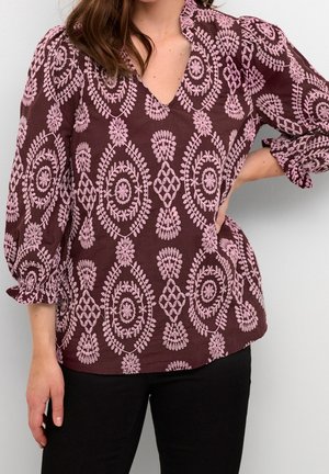 Kvinde iført en rødbrun bluse med lyserøde broderede blomster- og geometriske mønstre samt puffede trekvartærmer, kombineret med sorte bukser.