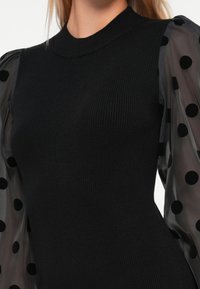 Abito nero a coste con maniche corte trasparenti a sbuffo caratterizzate da grandi pois neri. Scollo rotondo semplice e silhouette aderente.