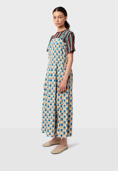 Pitkä kuvioitu mekko, jossa on sinisiä ja oransseja ympyröitä beige-taustalla, yhdistettynä vihreään ja punaiseen raidalliseen lyhythihaiseen paitaan.