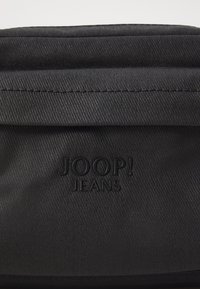 Svart tygväska med en slät textur, prydd med en embossed "JOOP! JEANS"-logotyp. Designen inkluderar sydda kanter och en platt, rektangulär form.