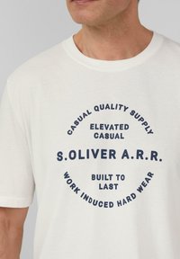 Korte mouw, ronde hals T-shirt van lichtcrème katoen met marineblauwe grafische tekst: "S.OLIVER A.R.R." en zinnen die het midden omcirkelen.