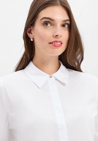 Jonge vrouw met lang bruin haar, gekleed in zilveren oorbellen en een witte blouse met kraag, die iets naar rechts kijkt op een witte achtergrond.