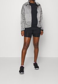 Giacca leggera zip-up argento e nera con cappuccio; t-shirt nera e pantaloni corti neri; abbinata a scarpe sportive nere.