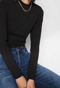 Schwarzes langärmliges Oberteil aus weichem Stoff mit hohem Halsansatz, kombiniert mit einer silbernen Kettenhalskette. Dazu werden blaue Jeans getragen.