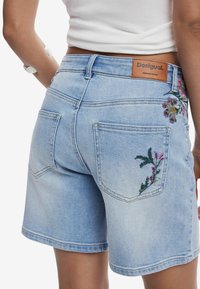 Pantalones cortos de mezclilla azul claro con bordado floral en los bolsillos traseros y una etiqueta de marca de cuero, con un ajuste estructurado y un diseño casual.