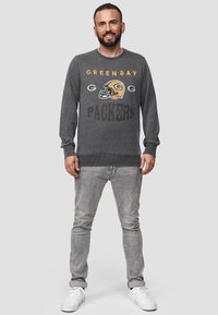 Felpa grigia con grafica "GREENBAY PACKERS" e design del casco; polsini e orlo a coste, abbinata a jeans grigi chiari e sneakers bianche.