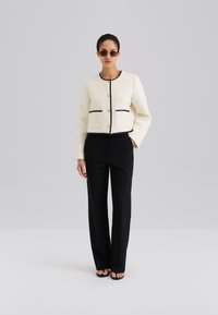 Biała krótka kurtka z czarnymi akcentami i złotymi guzikami, w zestawie z czarnymi szerokimi spodniami i czarnymi klapkami. Modelka nosi ciemne okulary przeciwsłoneczne.