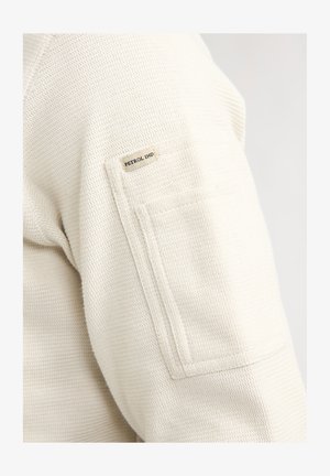 Primer plano de la manga de una camisa de tela texturizada beige con un bolsillo rectangular y una pequeña etiqueta "Petrol Ind" encima del bolsillo.
