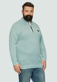 Jasnoniebieski sweter z fakturowanej tkaniny, z kołnierzem na pół zamku i małym logo na klatce piersiowej, noszony z niebieskimi dżinsami.