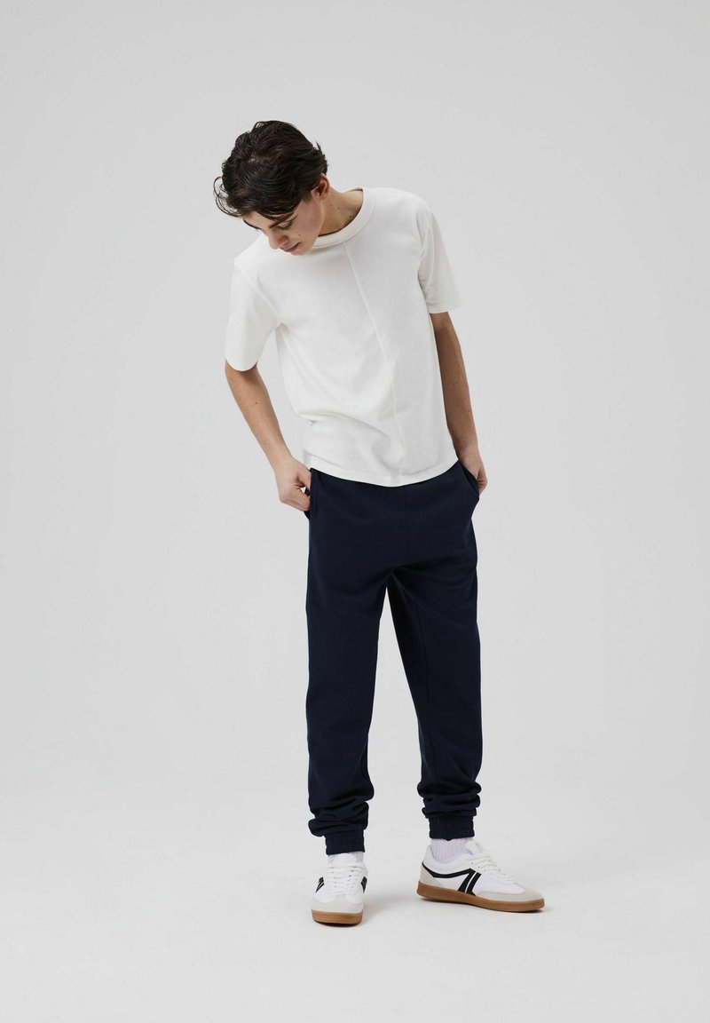Weißes, kurzärmliges Hemd, lockere marineblaue Jogginghose und weiße Sneakers mit schwarzen Akzenten. Einfaches Design, weicher Stoff und lässige Passform.