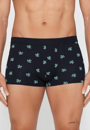 Männlicher Oberkörper trägt marineblaue Boxershorts mit hellblauem Schildkrötenmuster, steht mit entspannten Händen an den Seiten.