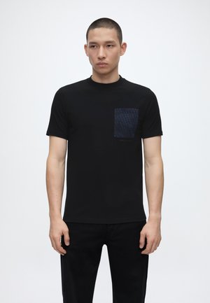 Mann iført en svart kortermet t-skjorte med en mørk mønstret brystlomme, stående foran en ensfarget hvit bakgrunn.