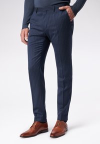 Marynarskie spodnie w prążki w dopasowanym kroju, wykonane z gładkiego materiału, z płaskim przodem i bocznymi kieszeniami. Brązowe skórzane eleganckie buty dopełniają całość.