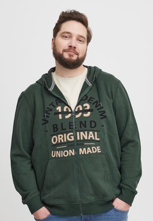 Green zip-up hoodie made of soft fabric, featuring a large front print with text in black and beige. Includes a drawstring hood and front pockets.