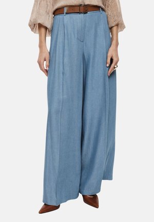 Donna che indossa pantaloni azzurri a vita alta e gamba larga, cintura marrone, camicetta beige, scarpe marroni con punta e un anello vistoso sulla mano destra.