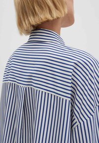 Gestreifte Bluse mit vertikalen blau-weißen Streifen, hohem Kragen und lockerem Schnitt. Leichter Stoff mit glatter Textur.