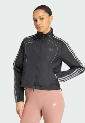 Frau trägt eine schwarze Adidas Zip-Up-Jacke mit weißen Streifen und hellrosa Leggings, steht mit einer Hand in der Tasche.