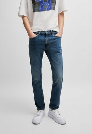 Person trägt ein weißes T-Shirt mit Grafik, blaue Slim-Fit-Jeans und weiße Sneaker, steht mit den Händen in den Taschen.