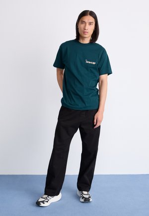 Teal bomull t-shirt med en liten grafisk print, ihop med lösa svarta byxor och svart-vita sneakers på ett blått golv.