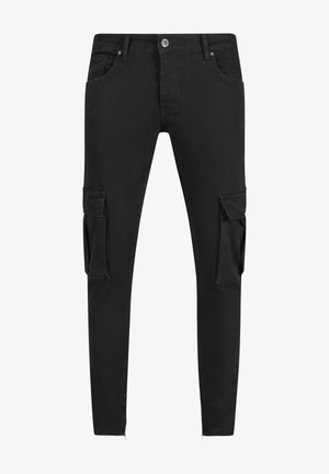 Mężczyzna w czarnej koszulce bez rękawów, czarnych spodniach typu cargo i czarno-białych tenisówkach stoi na szarym tle.