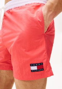 Păshorturi roșii de înot dintr-un material ușor, dotate cu un talie elastică albă cu șnur și o etichetă neagră inscripționată cu "TOMMY HILFIGER."