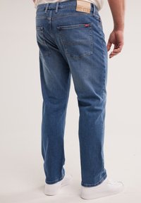 Férfi kék, egyenes szárú farmerben és fehér tornacipőben, hátulnézetben, egyszerű háttér előtt.