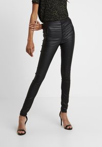 Pantalon noir en simili cuir à taille haute, avec des boutons et des poches latérales. Porté avec un haut à motif et des sandales noires à talons hauts.