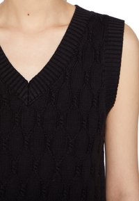 Close-up van een persoon die een zwarte, kabelgebreide mouwloze trui met een geribde V-hals draagt.