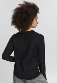 Melnā adīta džemperis ar apaļo apkakli, garām piedurknēm un brīvu piegriezumu. Tekstūra izskatās mīksta un nedaudz pūkaina.