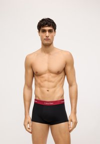 Čierne bavlnené boxer slipy s bordovým pásom s logom "Calvin Klein", predvádzané mužskou postavou s vyrysovanou postavou.