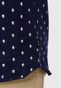 Camisa azul marino con patrón de diamantes blancos, de manga corta y con abertura lateral. Presenta una pequeña etiqueta negra con "Est. 1993." Tejido suave y texturizado.