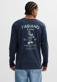 Hombre con cabello trenzado que lleva una camisa de manga larga azul marino con el logo del taller de reparación "Fabiano & Sons" y un personaje de llave inglesa de dibujos animados en la espalda, combinado con jeans.