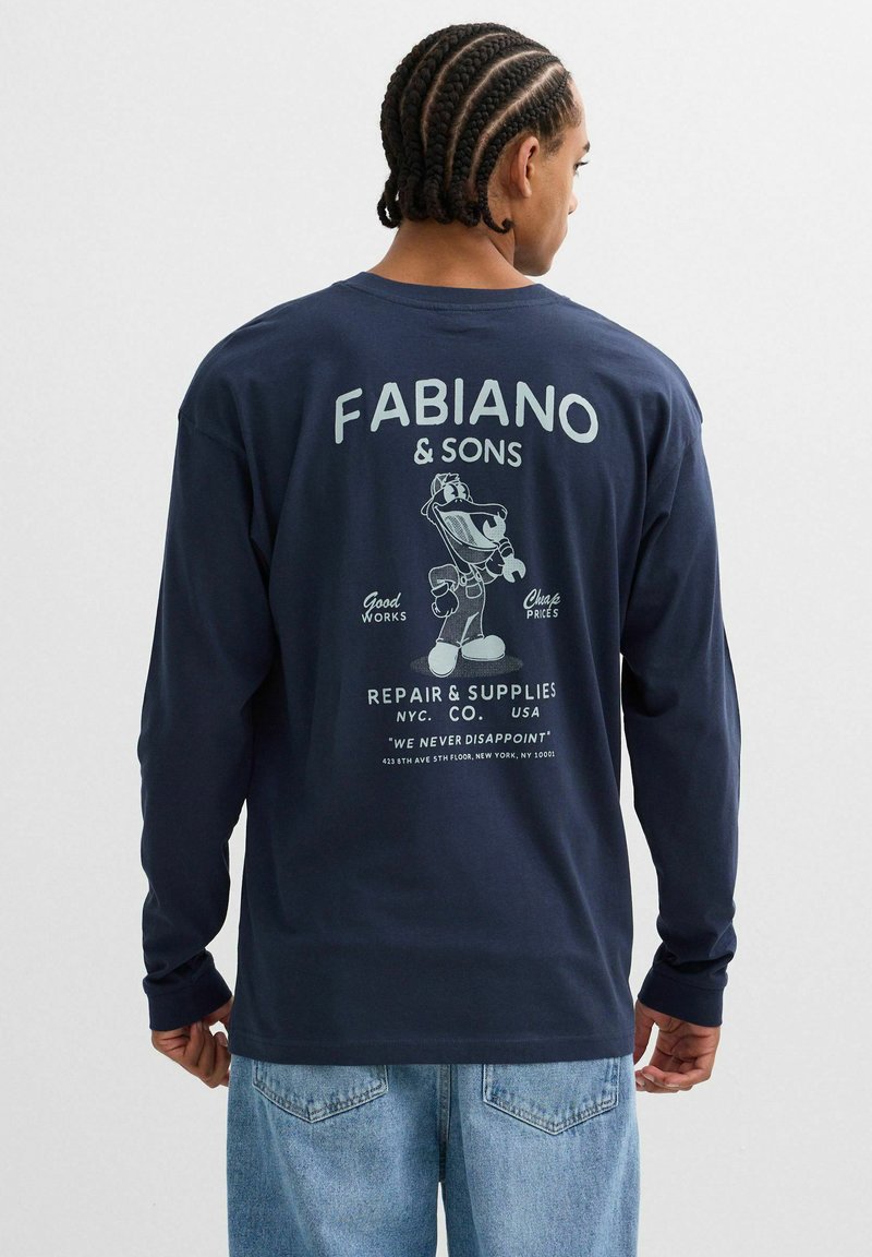 Hombre con cabello trenzado que lleva una camisa de manga larga azul marino con el logo del taller de reparación "Fabiano & Sons" y un personaje de llave inglesa de dibujos animados en la espalda, combinado con jeans.
