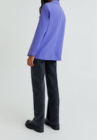 Persona che indossa un blazer lilla, jeans neri a gamba dritta con orli sfrangiati e scarpe chunky nere, in piedi di fronte a uno sfondo leggero e uniforme.