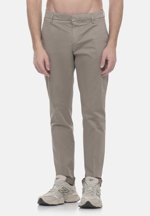 Uomo che indossa pantaloni beige slim-fit e sneaker New Balance grigio chiaro, in piedi contro uno sfondo bianco uniforme.