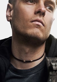 Svart flettet choker-halskjede med metalliske detaljer. Bært på en bar hals, kombinert med en mørk hettegenser og skinnjakke.
