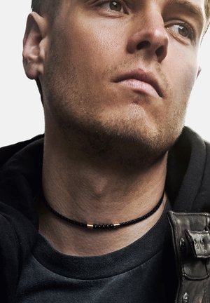 Svart flettet choker-halskjede med metalliske detaljer. Bært på en bar hals, kombinert med en mørk hettegenser og skinnjakke.