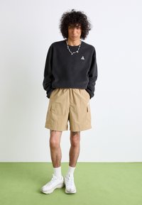 Czarna bluza z małym logo w kształcie trójkąta, w zestawieniu z beżowymi szortami cargo. Białe sneakersy i białe skarpetki do kostek dopełniają stylizację.