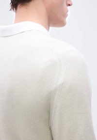 Gros plan sur le dos et le profil latéral d'une personne portant un pull en maille texturée de couleur crème avec un col blanc, sur un fond uni.