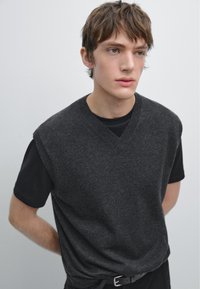 Jonge man met kort bruin haar, gekleed in een donkergrijze mouwloze sweater over een zwart T-shirt, staande met zijn handen achter zijn rug.