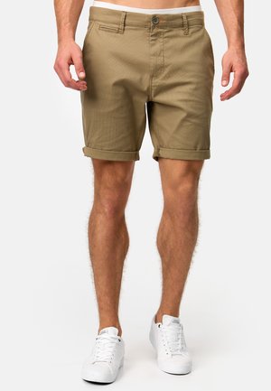 Beige Baumwollshorts mit umgeschlagenen Säumen, einem einzelnen Fronttasche, Gürtelschlaufen und einem vorderen Knopfverschluss. Getragen mit weißen Turnschuhen.
