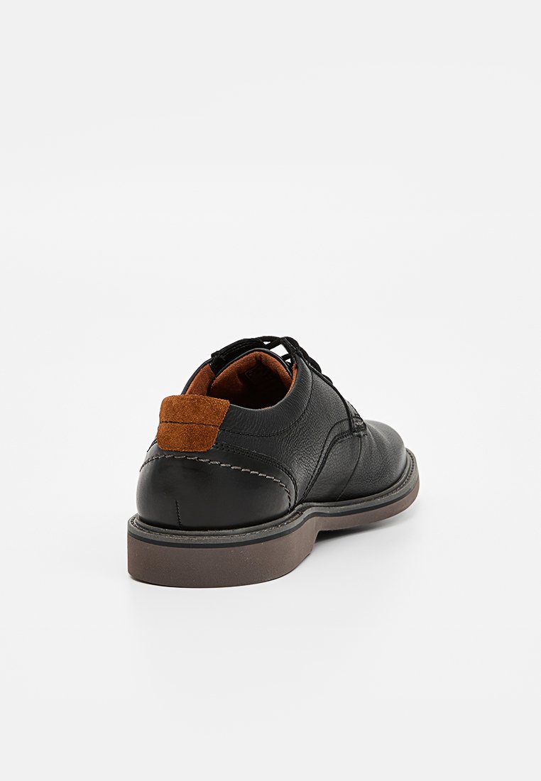 Scarpa in pelle nera con finitura liscia, caratterizzata da un accento in camoscio marrone sul tallone e una suola in gomma. Lacci visibili sulla parte superiore.