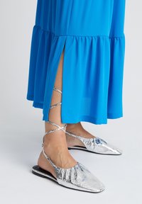 Chaussures plates à lacets en cuir métallisé argenté portées avec une robe bleue longueur cheville présentant une fente latérale sur fond blanc.