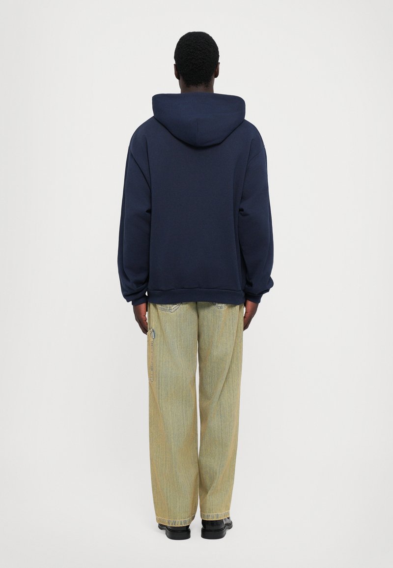 Navy hoodie with a large hood, relaxed fit, paired with light yellow wide-leg jeans featuring a subtle striped pattern and zippers.