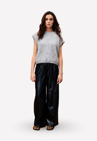 Vrouw staand, gekleed in een grijze donzige mouwloze top, wijde zwarte leren broek en schoenen met luipaardprint, tegen een plain witte achtergrond.