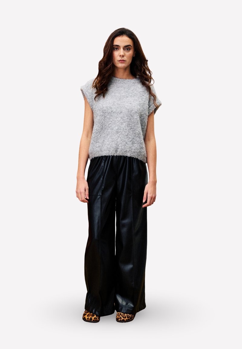 Femme debout, portant un haut sans manches gris duveteux, un large pantalon en cuir noir et des chaussures à imprimé léopard, contre un fond blanc uni.