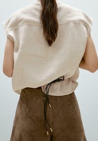 Jasno beżowy lniany top z krótkimi rękawami, zapinany z tyłu na czarne sznurki z metalicznymi akcentami. W połączeniu z brązowymi spodniami z zamszu.