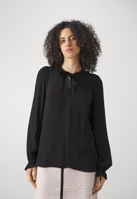 Blouse noire avec des manches bouffantes et un col à nouer. Tissu doux, texture lisse et léger froncé aux poignets.